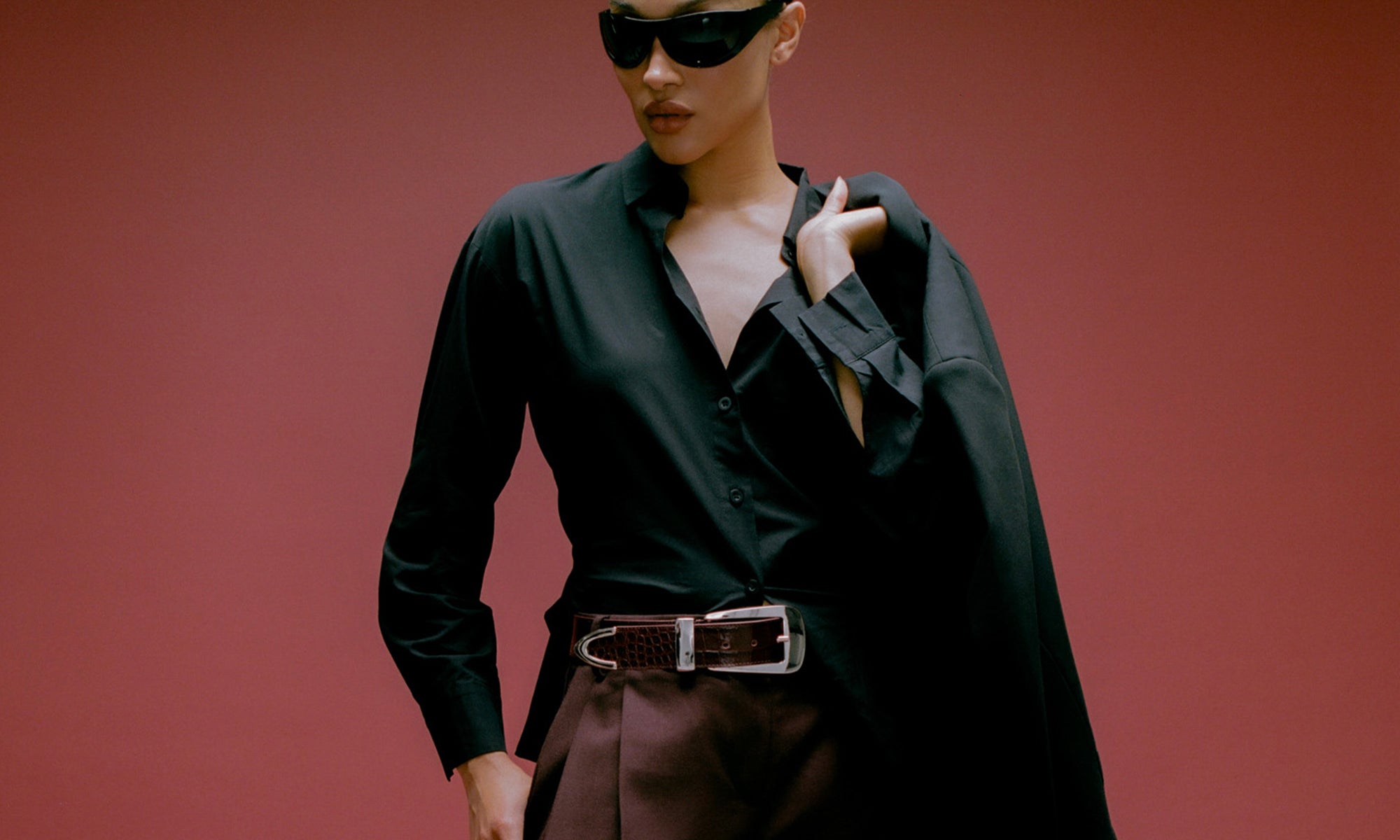 Person wearing a black outfit with sunglasses against a red background wearing burgundy belt with big silver buckle