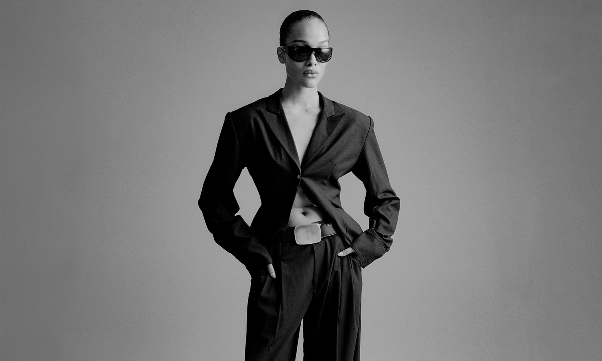 Person wearing a black suit and sunglasses on a gray background with big buckle belt