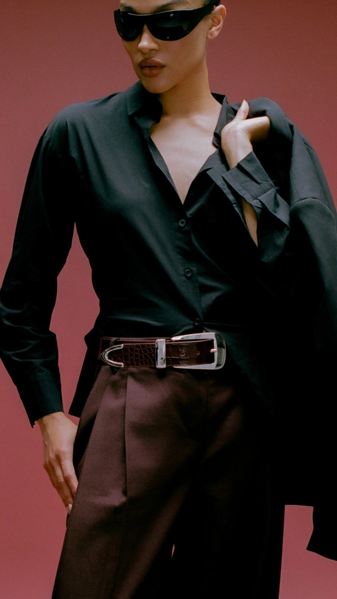 person wearing burgundy belt with big silver buckle. standing against a red backdrop with a black top and burgundy pants.