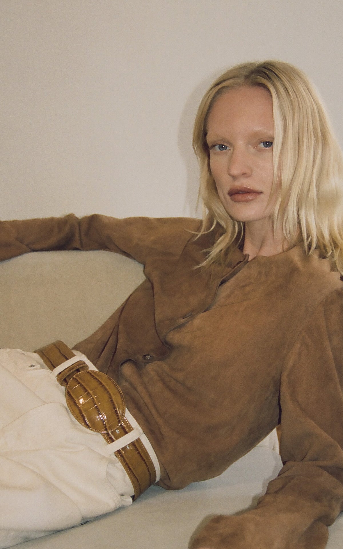 Woman wearing a brown suede jacket sitting on a white couch. Wearing a tan croc belt.