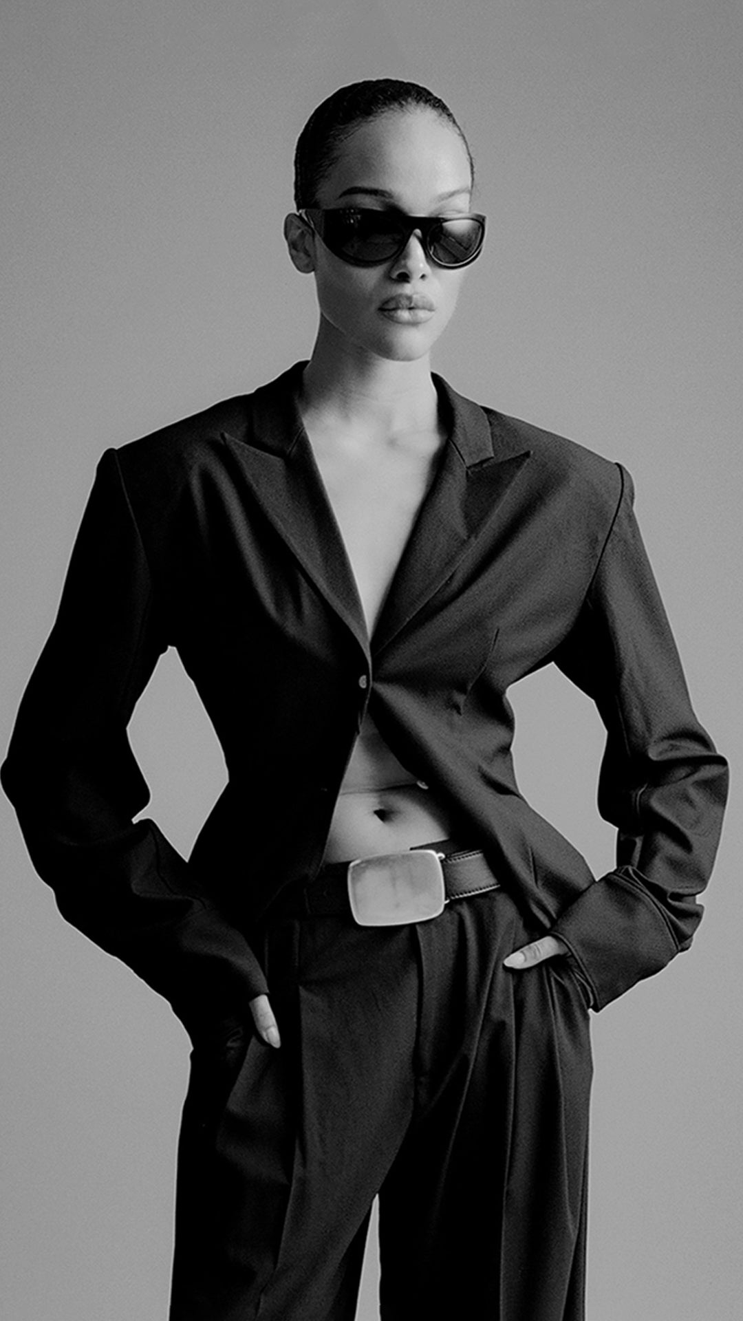 Person wearing a black suit and sunglasses against a gray background with big buckle belt