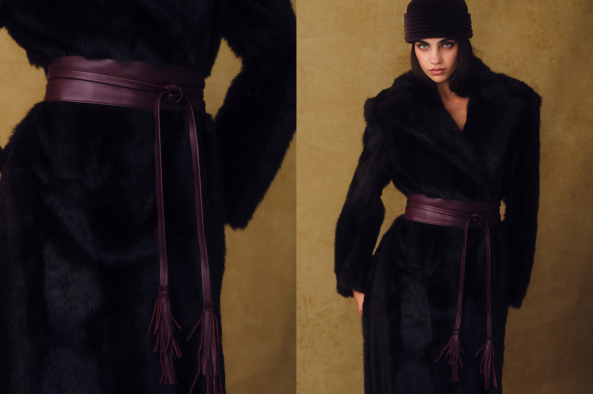 model against a yellow textured backdrop. wearing a dark fur coat and hat with a burgundy colored wide leather wrap belt.