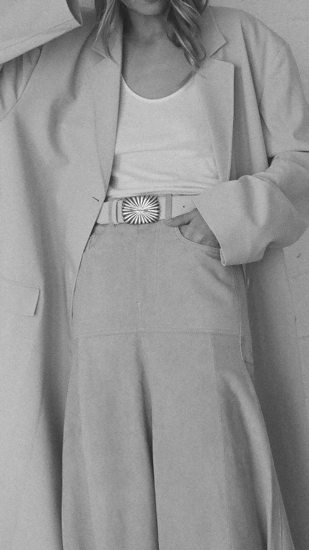 Person wearing a long coat with a belt and white top, in grayscale.