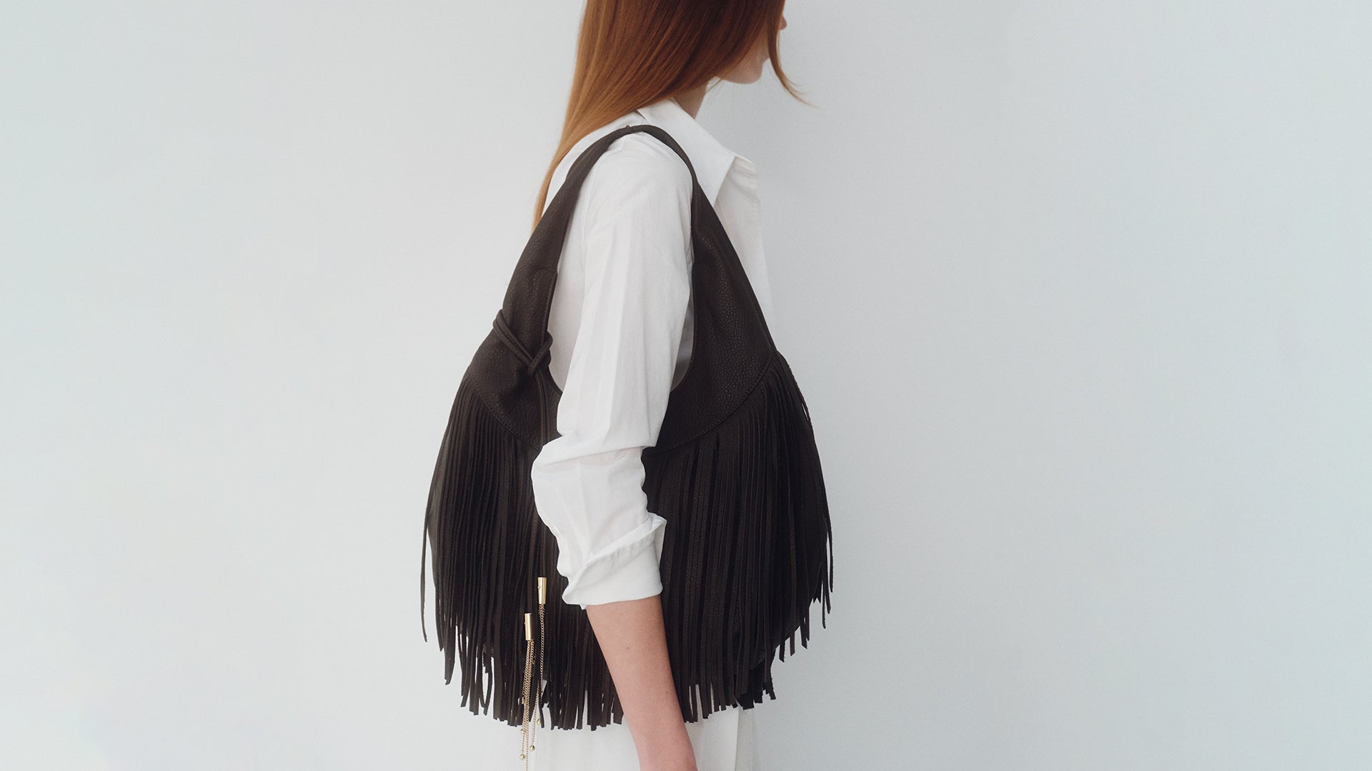 Person wearing a brown fringe hobo bag over a white shirt on a plain background