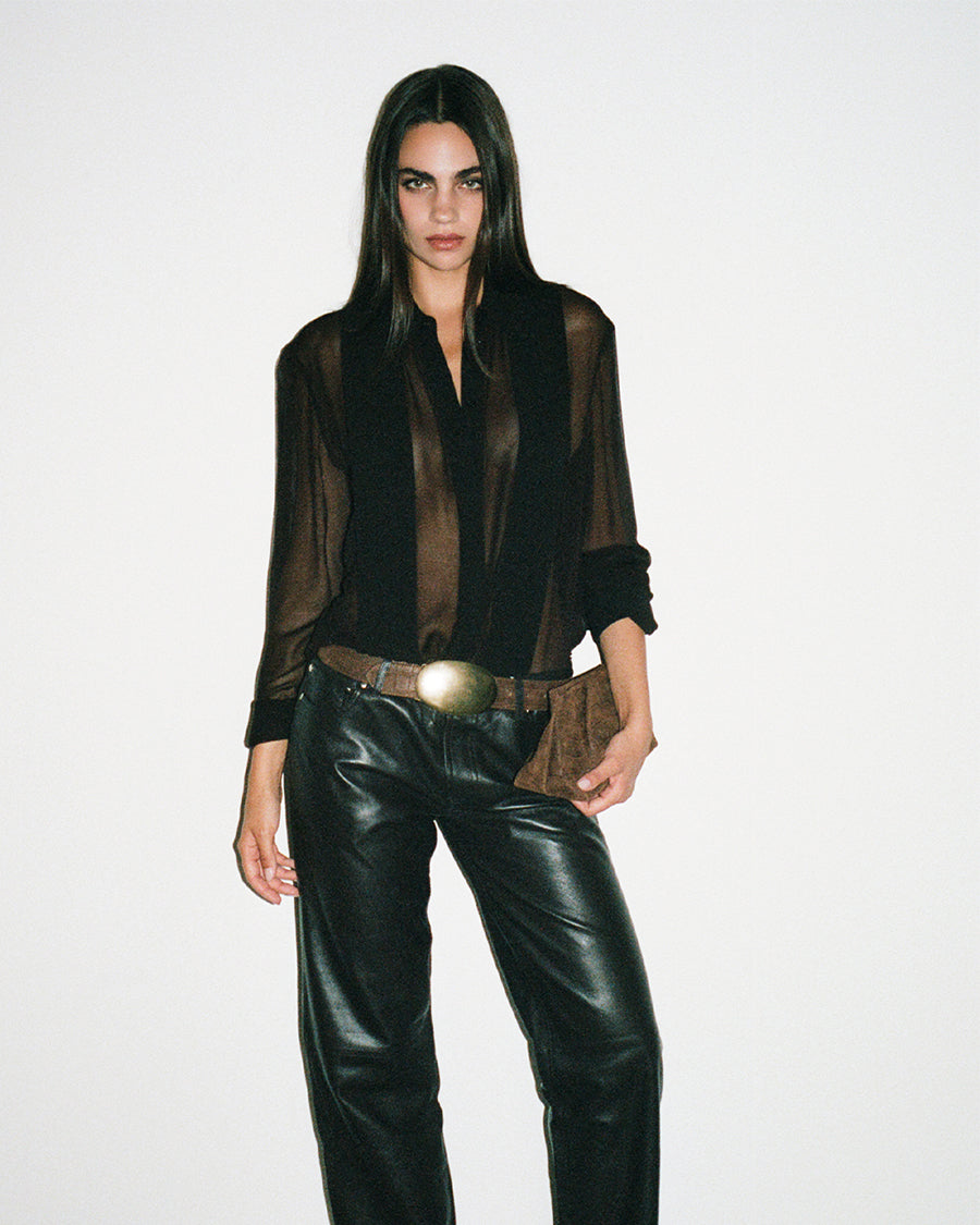 A model wearing a sheer black long-sleeve button-up shirt with a brown lining, paired with black leather pants and a brown belt with a large gold buckle, against a plain white studio background.