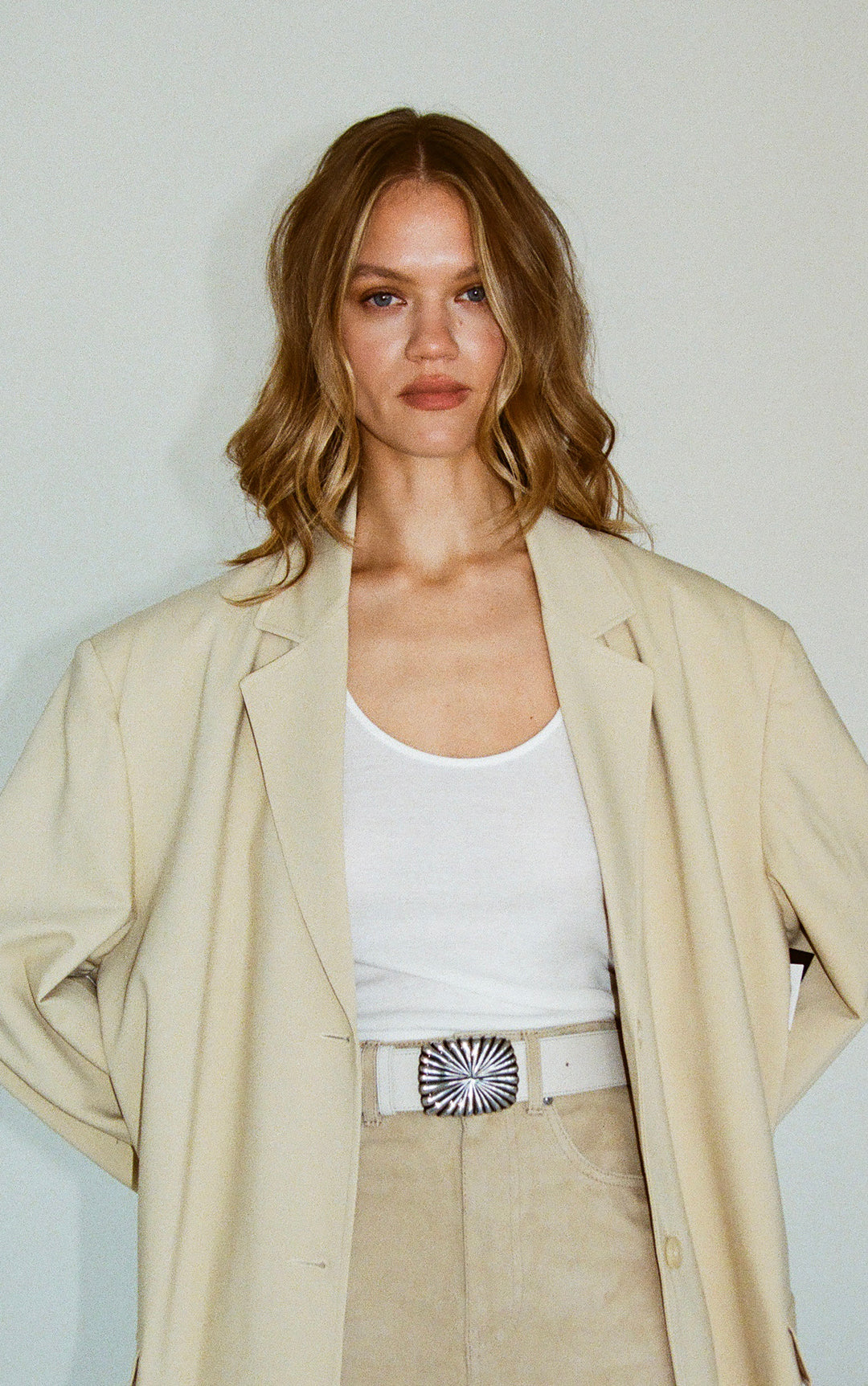 Woman wearing a beige blazer over a white top with a belt, against a plain background