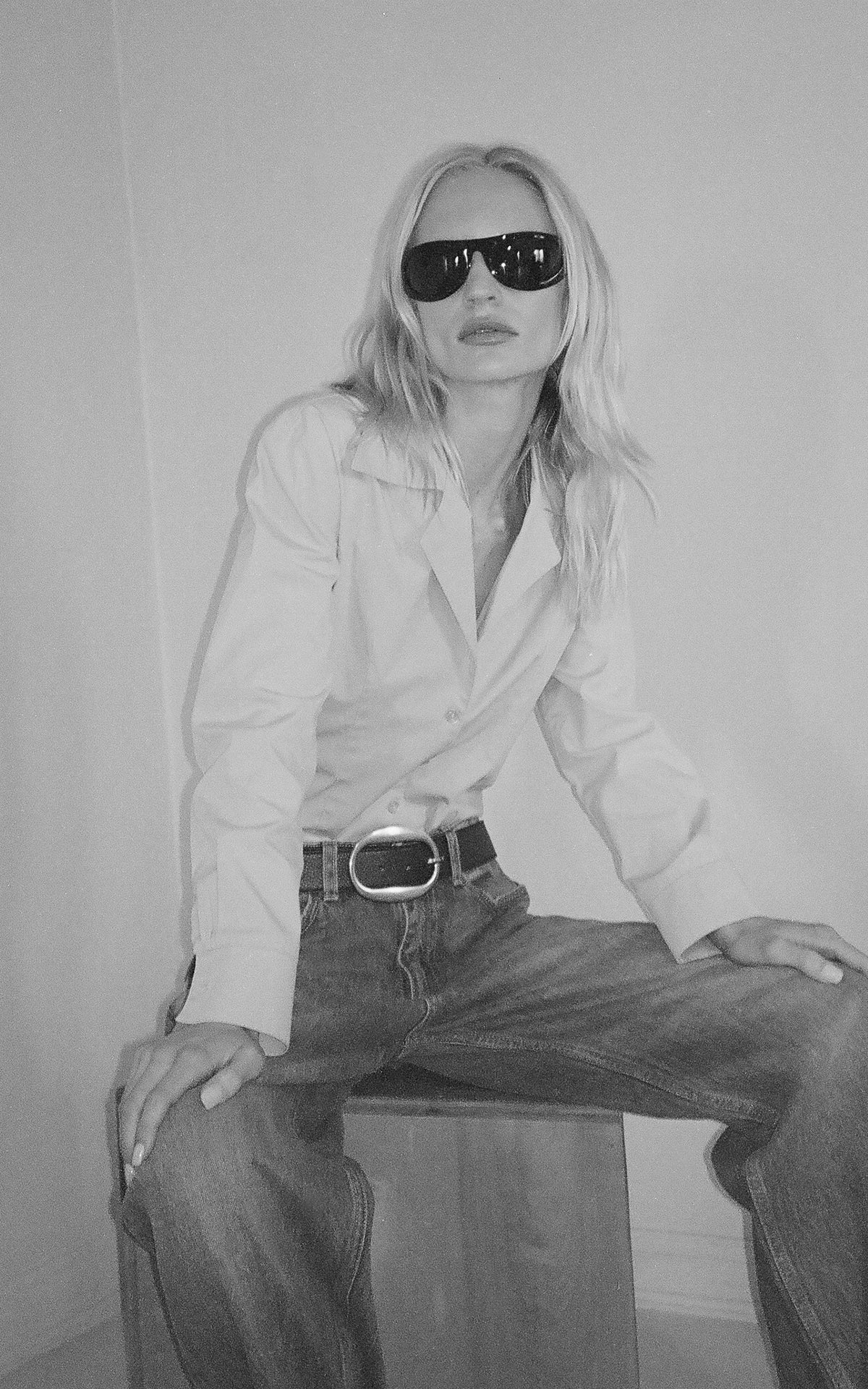 Person wearing sunglasses and a white shirt sitting on a stool against a plain background. Wearing a black leather belt with a brass oval buckle.