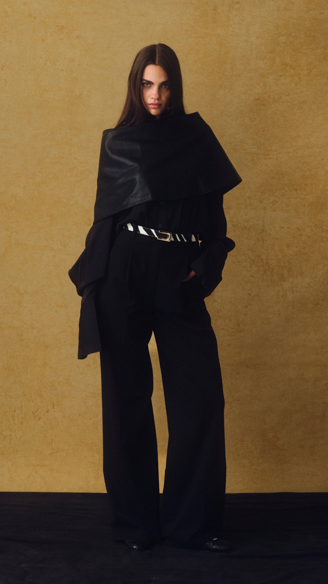 A model stands against a textured gold backdrop wearing a black wide-leg coat with a draped collar, black wide-leg trousers, and a black belt with silver hardware.