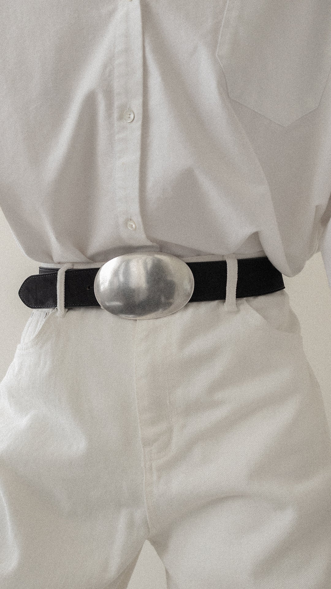 Person wearing a white shirt and white pants with a black belt featuring a large silver buckle.