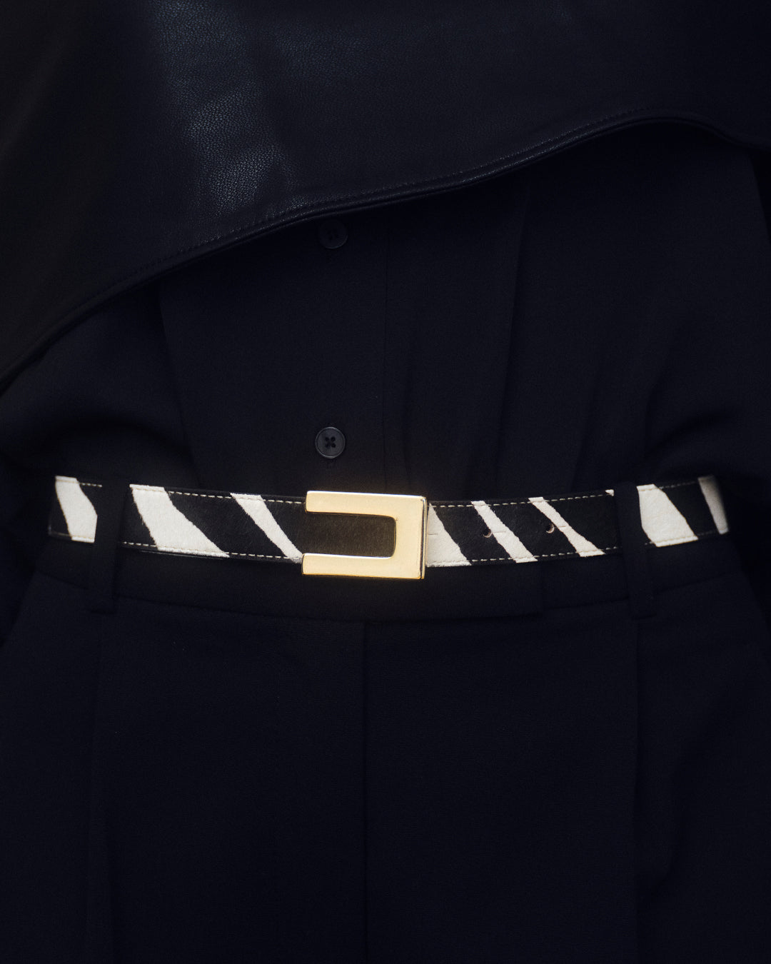 A close-up of a black and white zebra-patterned belt with a gold rectangular buckle, worn over a black shirt and trousers.