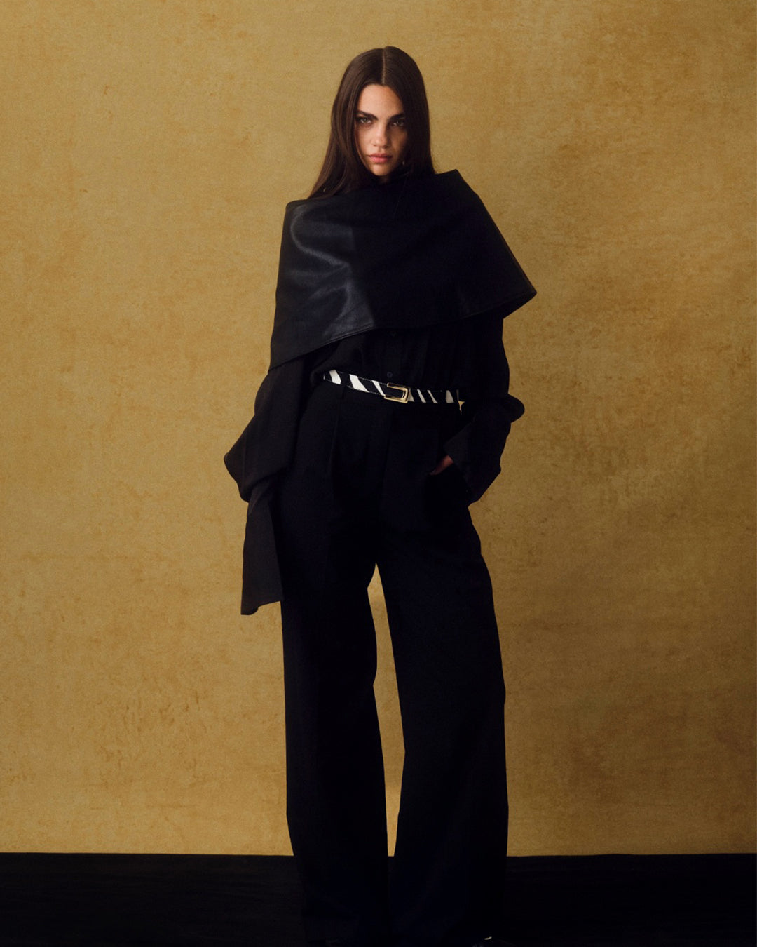 model wearing all black outfit against a textured yellow backdrop. Wearing a black and white zebra belt.