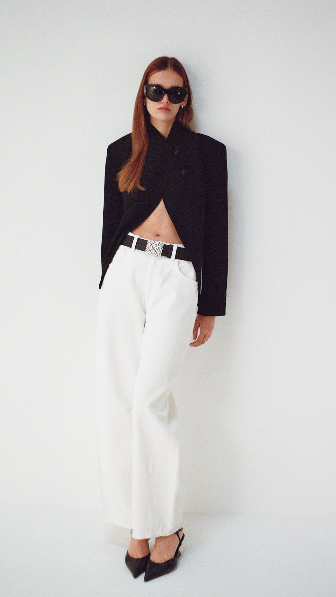 Woman wearing a black blazer and white pants on a white background wearing a black belt with a big silver buckle