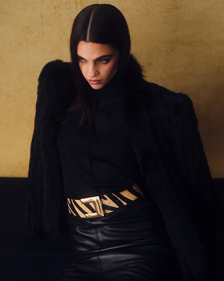 model wearing a wide tan zebra belt with oversized gold buckle on an all black outfit against a yellow backdrop